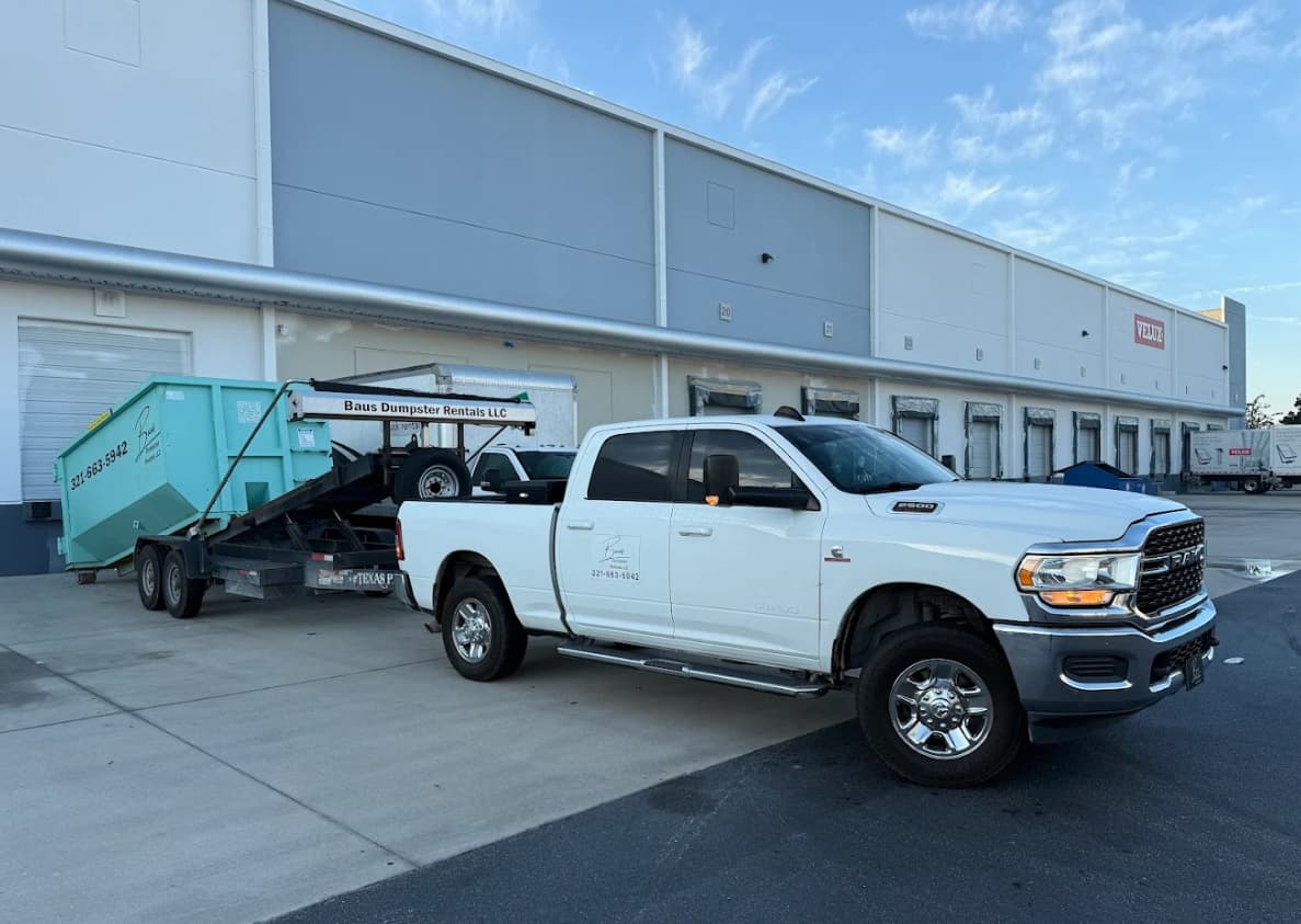 Baus truck towing a teal roll-off dumpster at a commercial warehouse in Kissimmee, FL