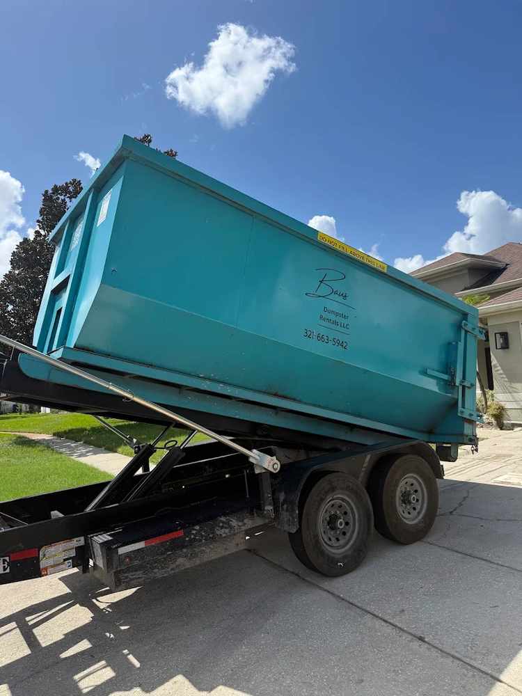 Baus Dumpster Rentals trailer unloading a teal roll-off dumpster at a home in Conway, FL